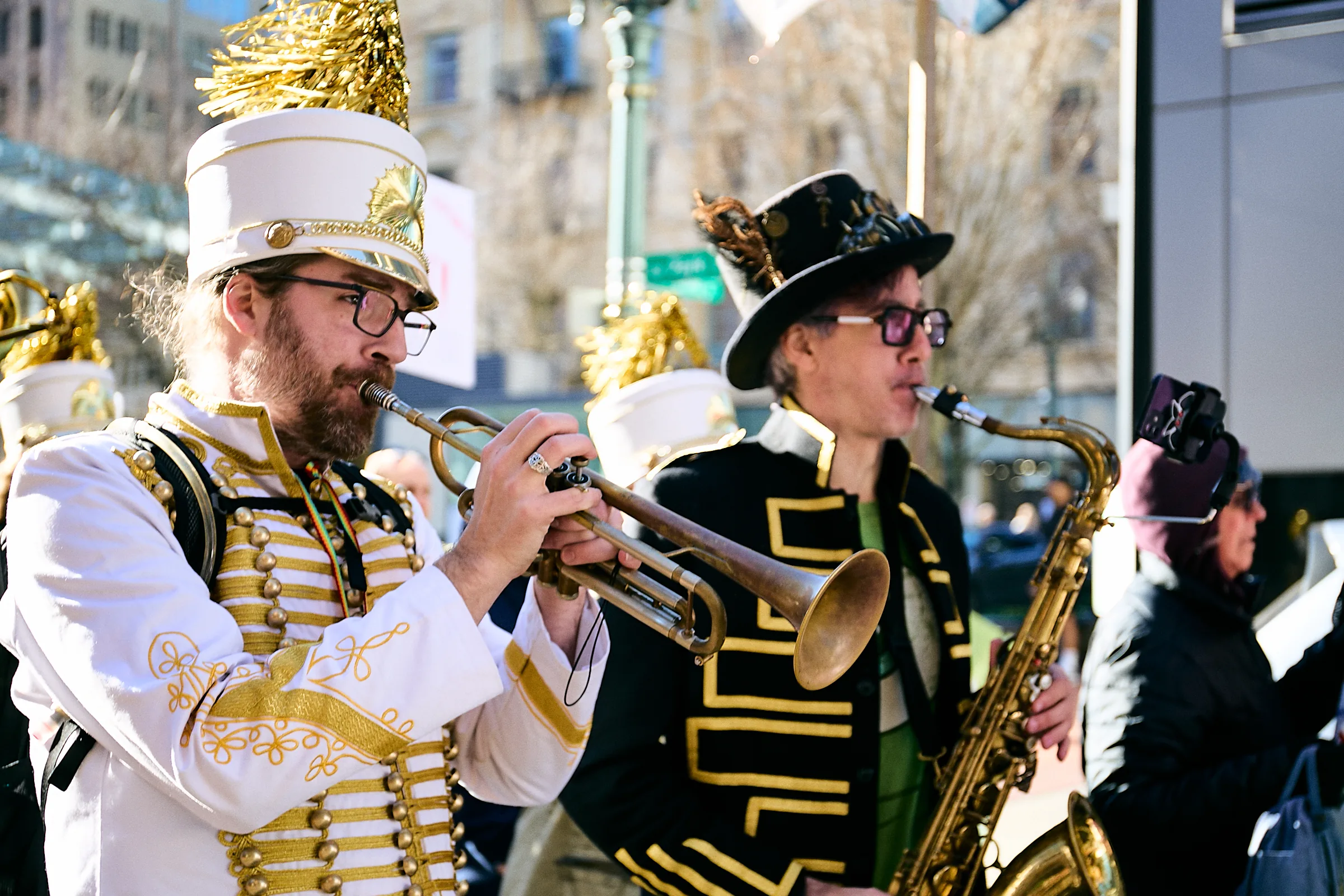 A trumpeter in a white-and-gold marching uniform and plumed shako and a saxophonist in a black top hat and black-and-gold jacket play side by side on a sunny downtown Portland street during the ICE Out of Portland march, January 17, 2026.